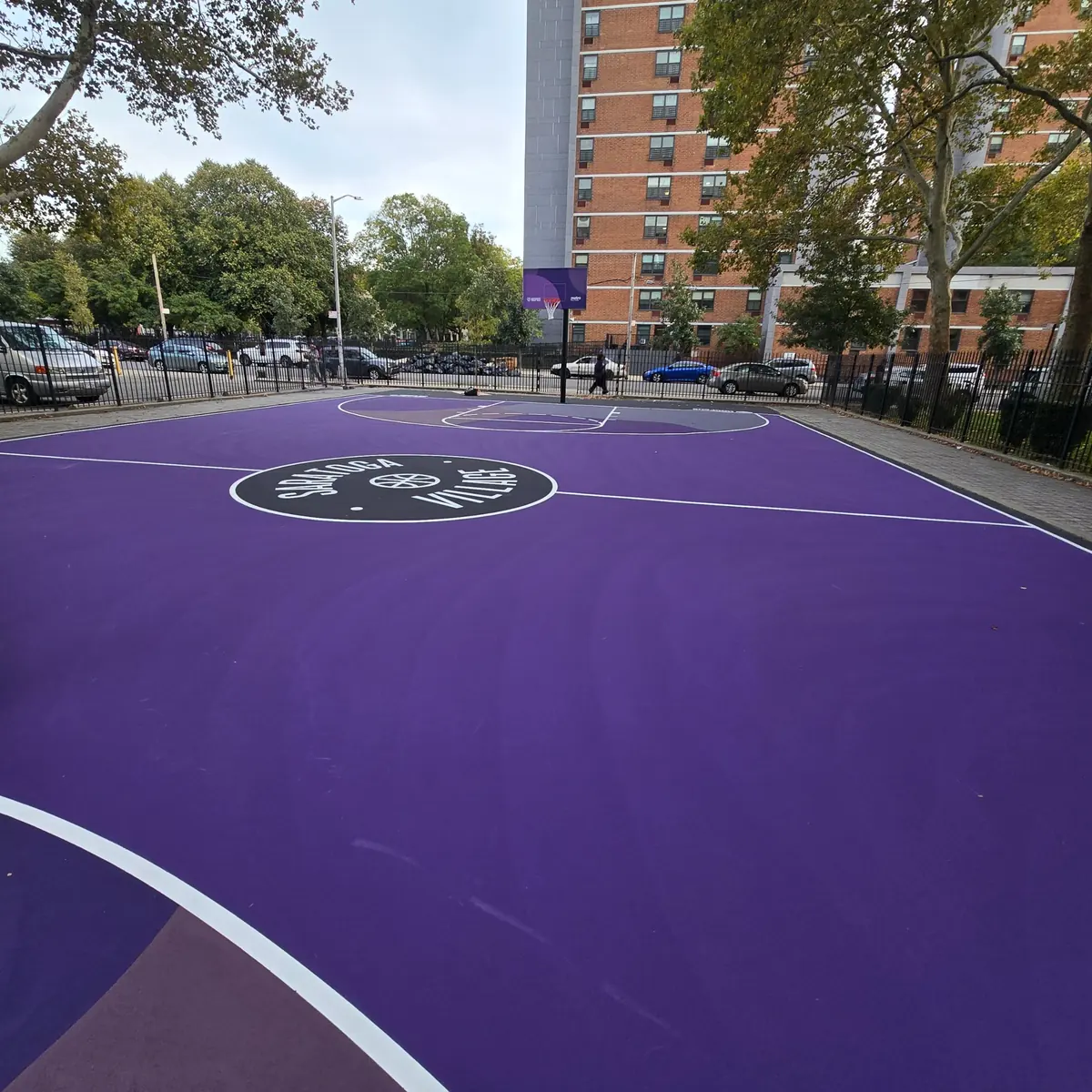 Ground-level view of the finished purple court at Saratoga Village, with the Saratoga Village badge at center court and a new backboard beyond