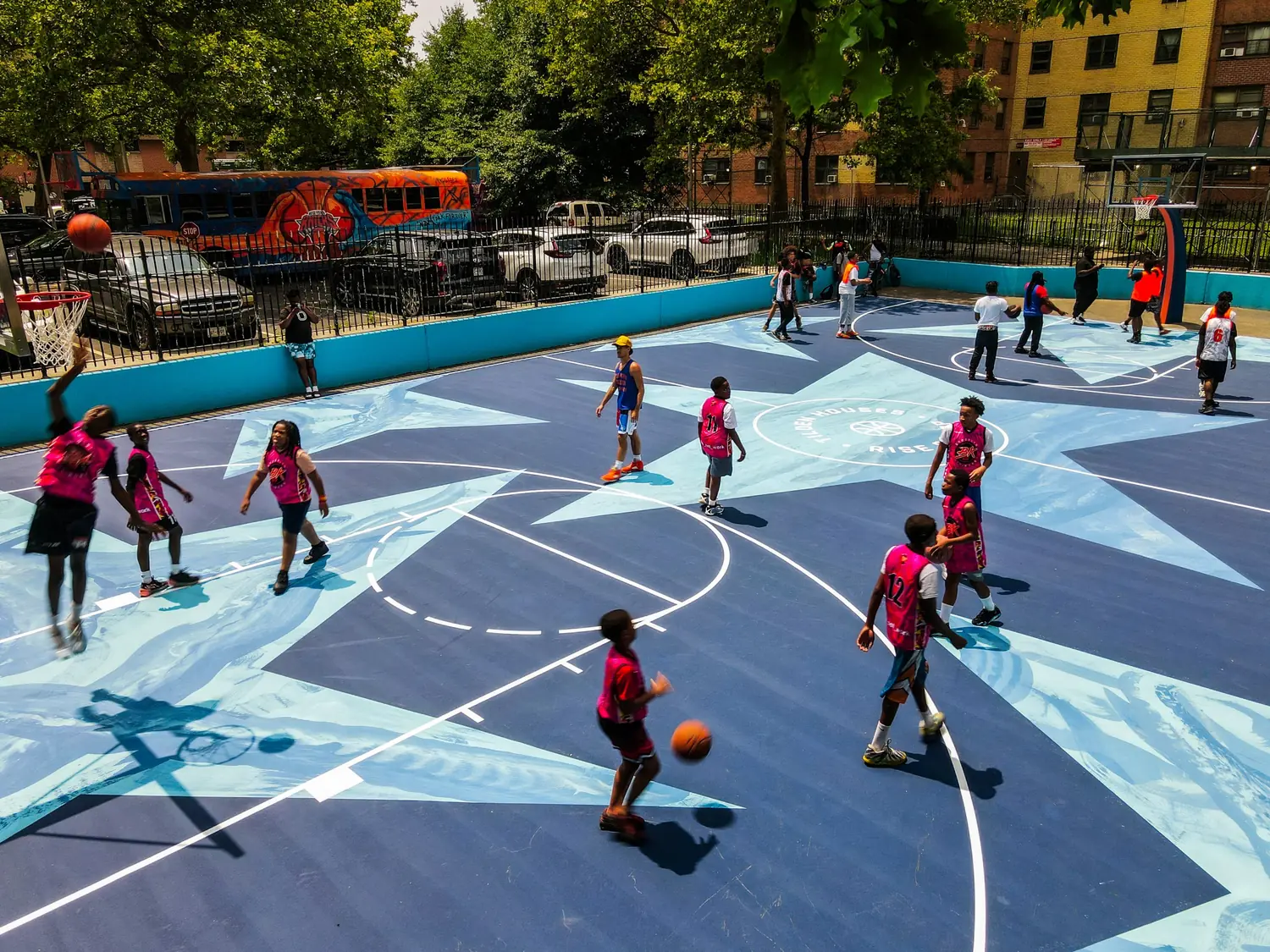 Young players in pink jerseys running a game on the finished Seven Stars Court, with the Hoopbus parked on the street behind the fence